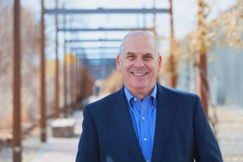 A man in a suit smiles in front of a fence.