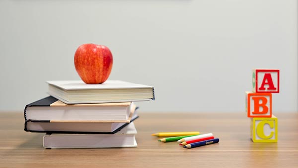 A stack of books with a red apple on top, a small group of colored pencils, and a stack of children's letter blocks all sit on top of a wooden surface.