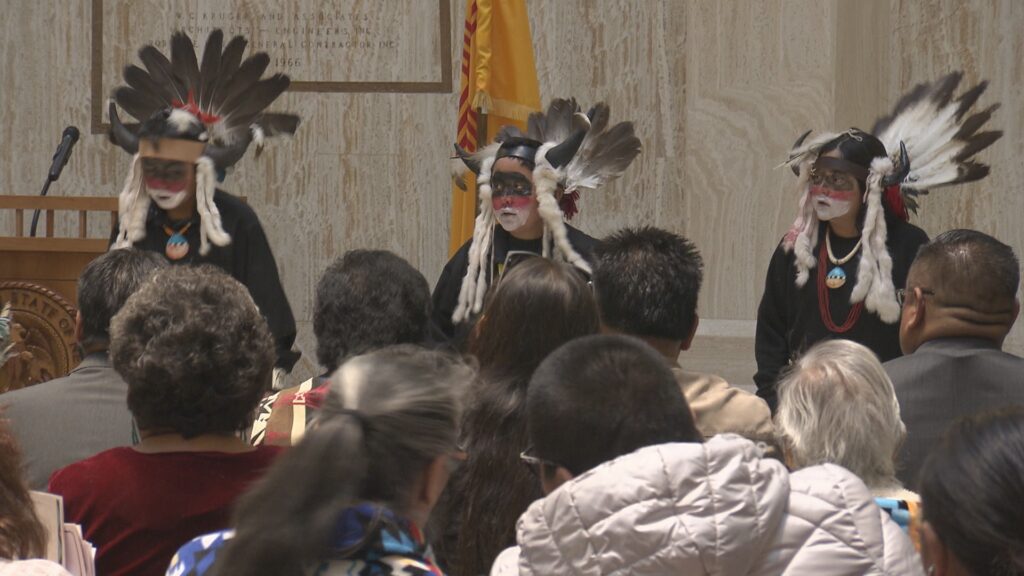A group of Indigenous dancers in traditional regalia performing in the Santa Fe Roundhouse.