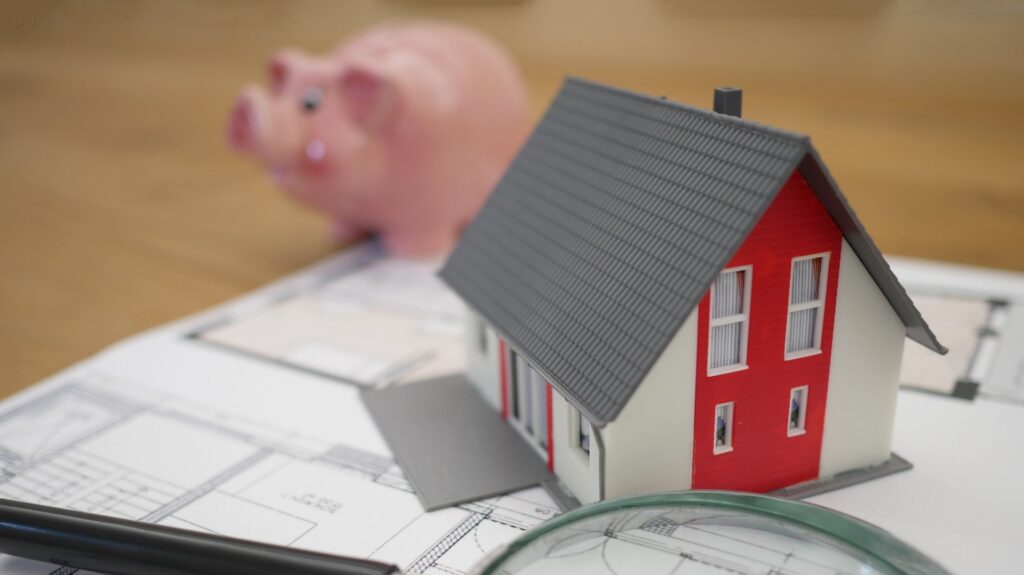 A model of a house sits on top of a blueprint, along with a pink piggy bank.