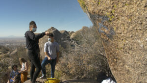 A group of masked people examine a rock structure in a desert area.