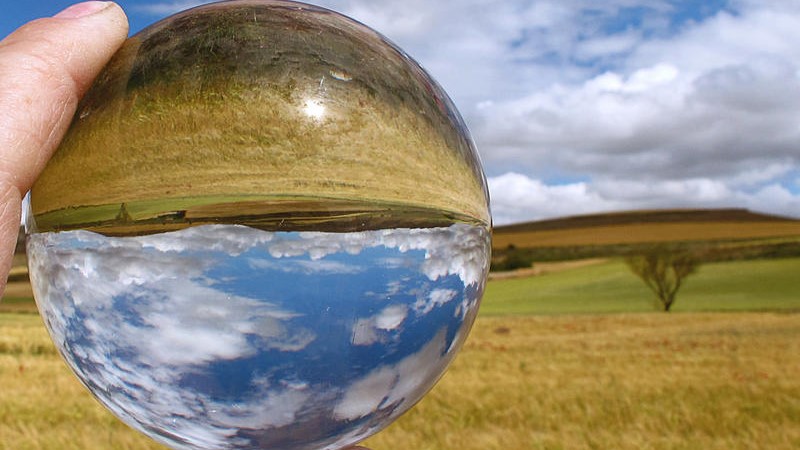 A hand holds a reflective sphere in the middle of a grassy plain.