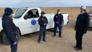 Four people in facemasks and gloves discuss with each other in a desert field.