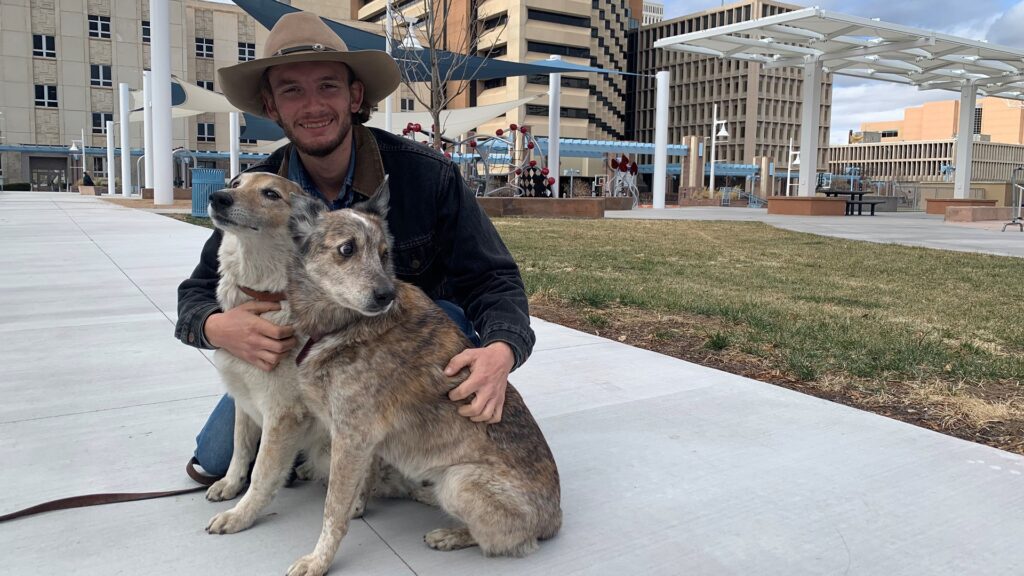 A person wearing a hat holds two dogs in a plaza.