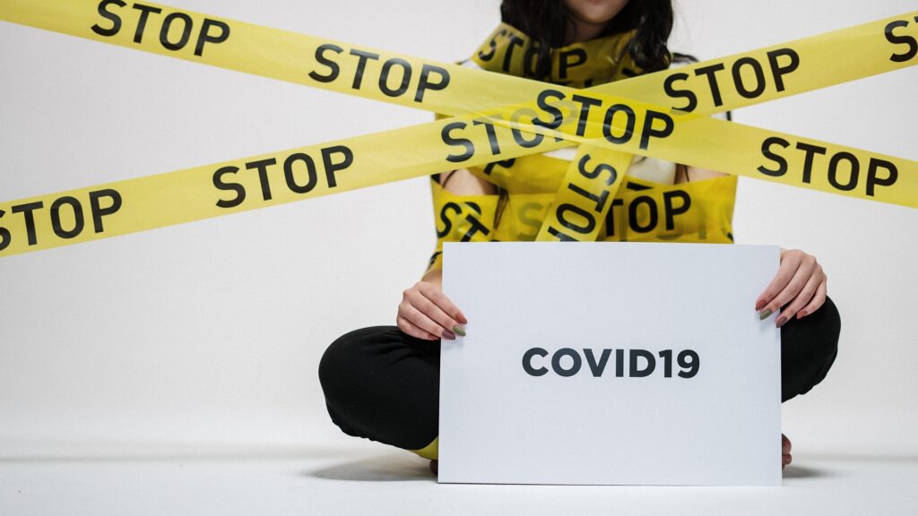 A person covered in crime scene tape that reads "STOP", and holding a sign that reads "COVID19".