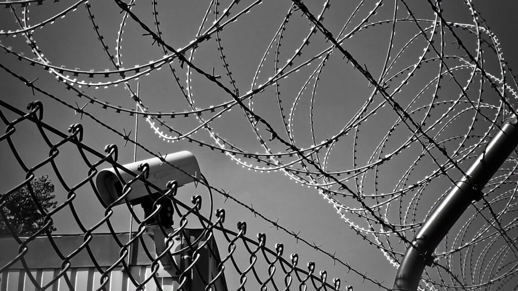Black-and-white image of a security camera looking down, with a fence and barbed wire in front of it.