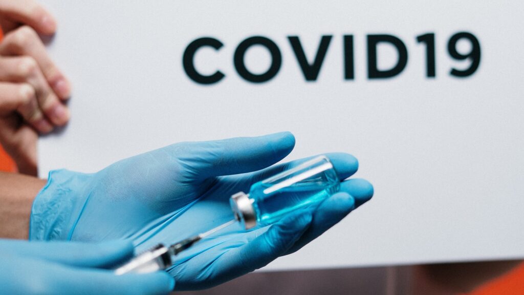 Gloved hands insert a syringe into a vial, with hands in the background holding a sign that reads "COVID19".