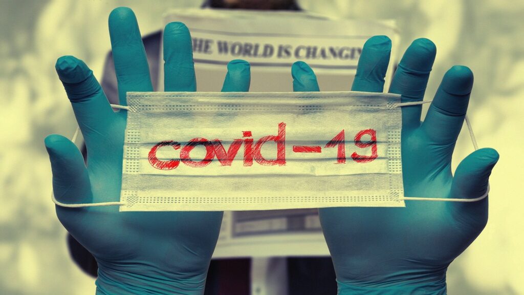 Two gloved hands hold a facemask reading "covid-19".