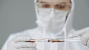 A person in a white bodysuit and wearing a facemask, holding a vial of dark liquid labeled "CORONAVIRUS".