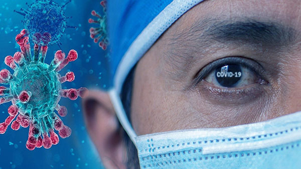 Composite of a nurse wearing a facemask and with "COVID-19" in their eye, with CG illustration of coronaviruses in the background.
