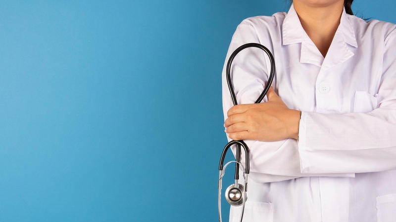 A faceless person in lab coat and holding a stethoscope in front of a blue backdrop.