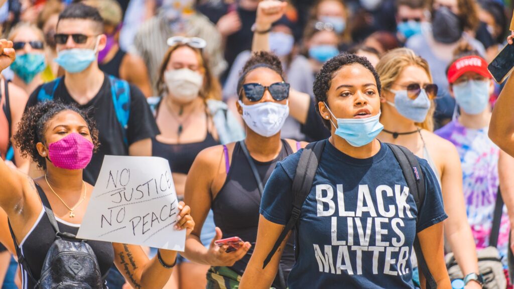 A group of demonstrators wearing facemasks, with one wearing a shirt reading "BLACK LIVES MATTER" and one holding a sign reading "NO JUSTICE NO PEACE".