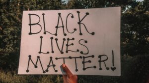 A hand holds up a sign reading "BLACK LIVES MATTER!"
