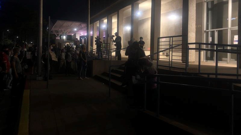 A group of demonstrators stand in front of Albuquerque City Hall at night.