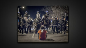 A person sits in the middle of a street as cops in riot gear, batons, and on horseback approach them.