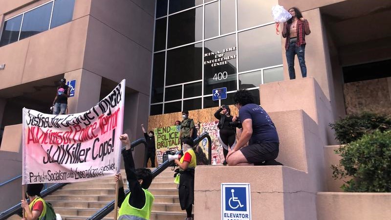 A group of demonstrators give speeches and hold signs in front of the Albuquerque Police Department during the day.