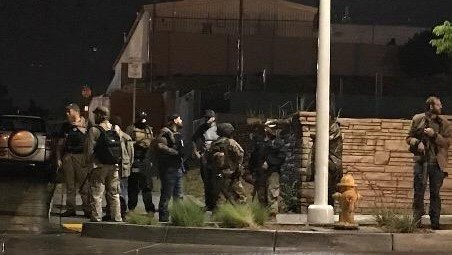 People in militia outfits and with guns stand on a sidewalk at night.
