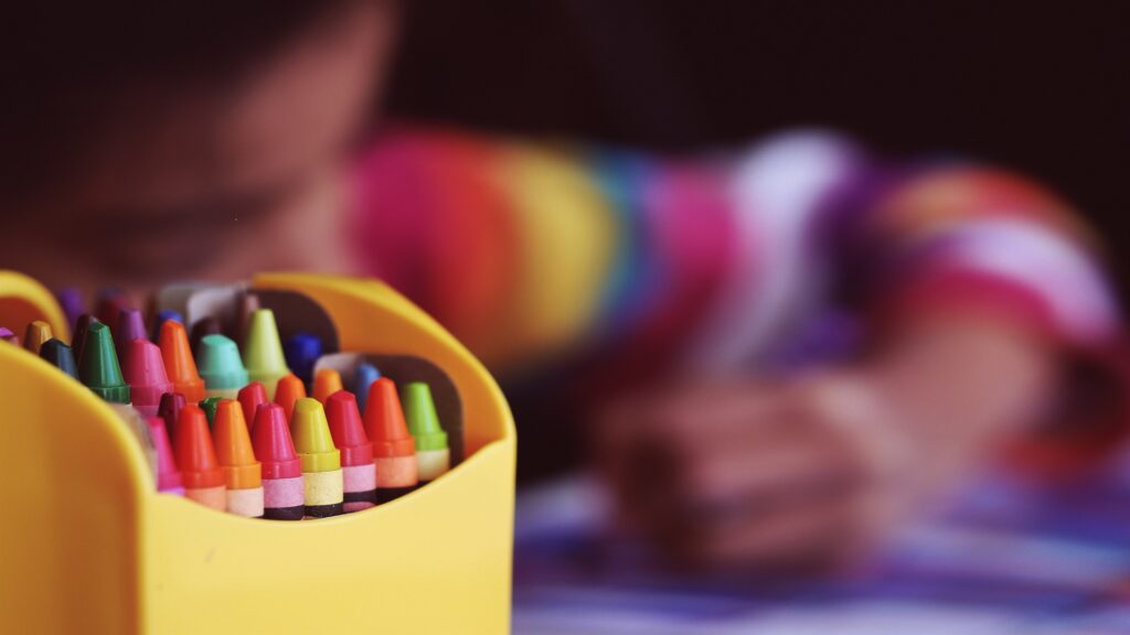 A box of crayons, with a child drawing in the background.