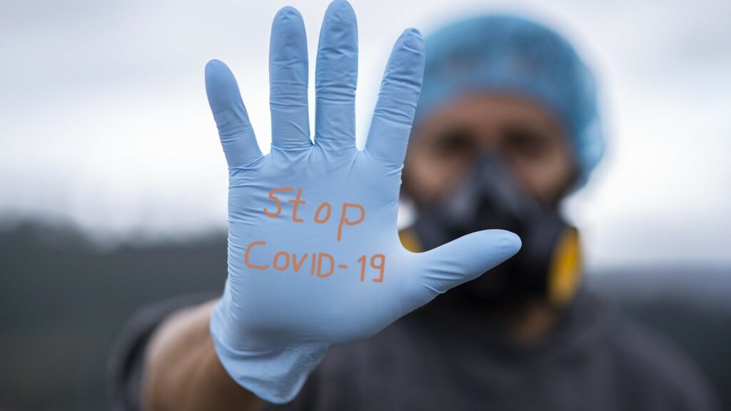 A person in hairnet and wearing a gas mask holds up a gloved hand that reads "Stop COVID-19".