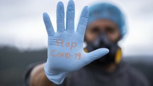 A person in hairnet and wearing a gas mask holds up a gloved hand that reads "Stop COVID-19".