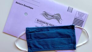 A mail ballot lies on a surface, with a cloth facemask lying on top of the ballot.