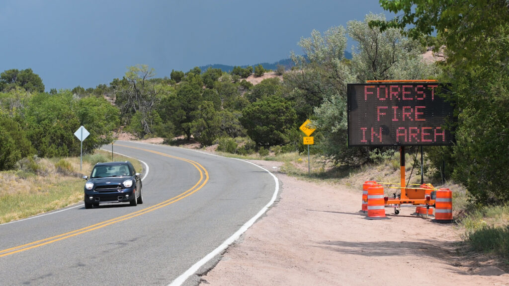 A car drives down a road that has a "FOREST FIRE IN AREA" sign posted next to it, against a dark blue sky.