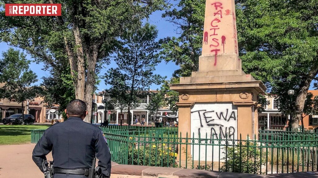 A cop stands in front of a vandalized obelisk, with graffiti reading "RACIST" and "TEWA LAND".