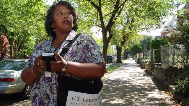 A Census worker looks up from their device on a sunny street.