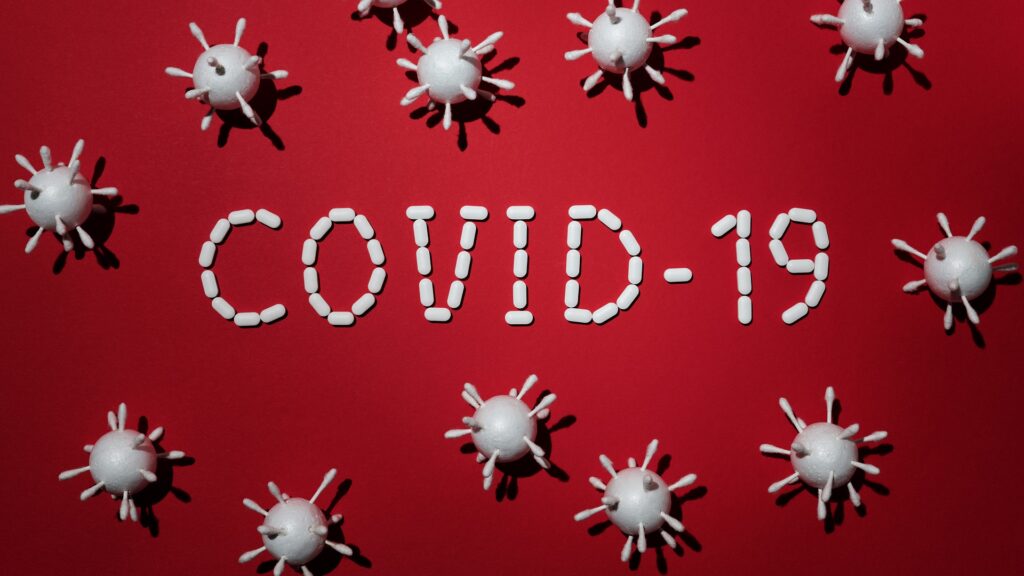 Coronaviruses surround a "COVID-19" made of pills and tablets, over a red backdrop.