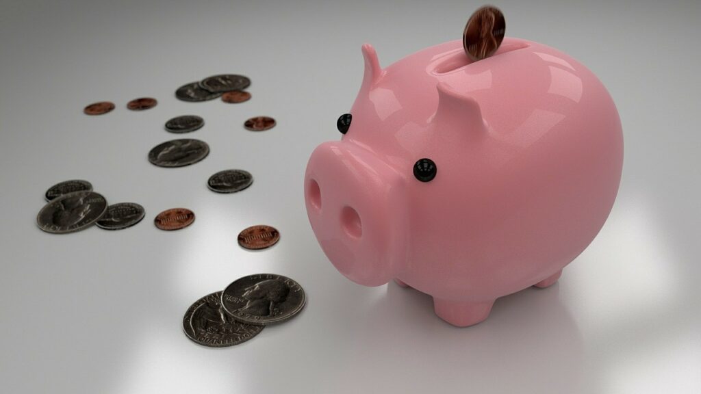 A piggy bank stands on a surface, with coins scattered across the surface.