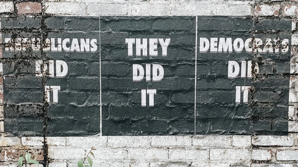 Signs printed on a brick wall reading "REPUBLICANS DID IT", "DEMOCRATS DID IT" and "THEY DID IT" in the center.