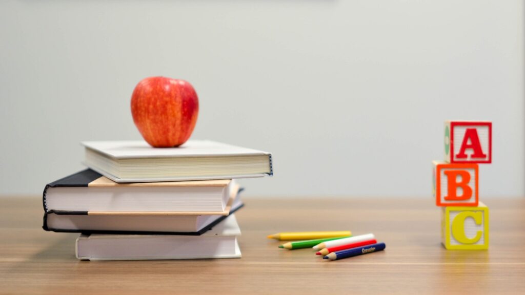 An apple, books, colored pencils, and letter blocks sit on top of a table.