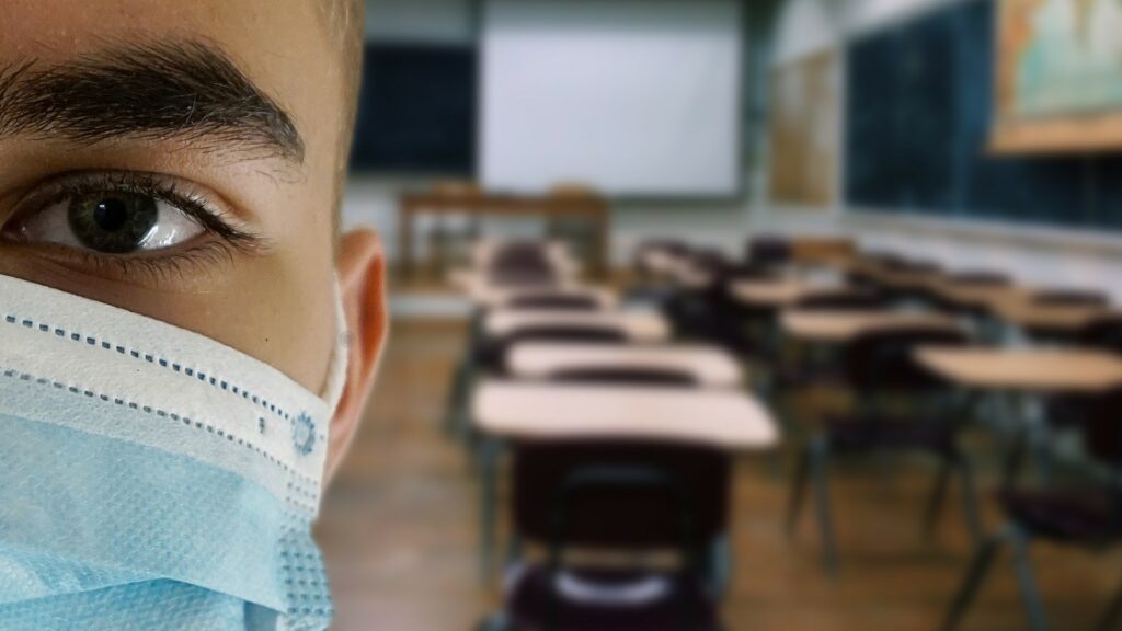 A person wearing a facemask, with an empty classroom behind them.