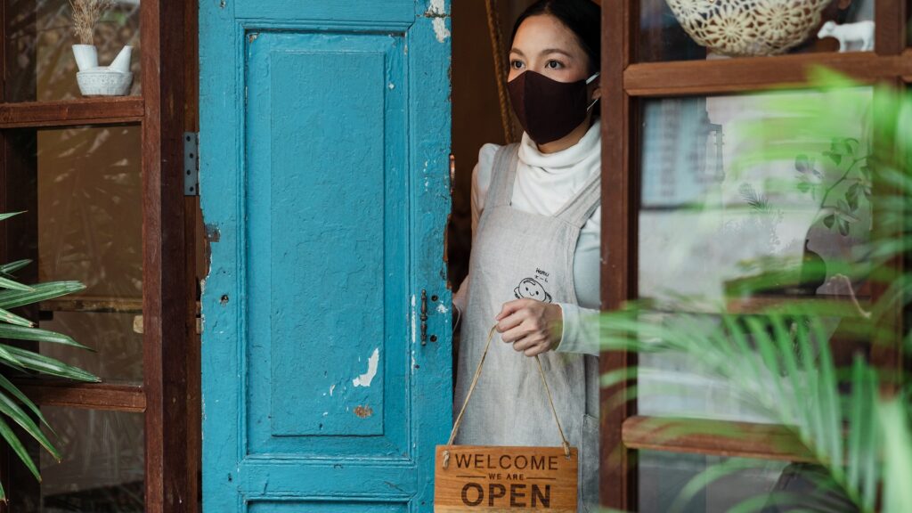 A person wearing a facemask holds a sign reading "WELCOME WE ARE OPEN".