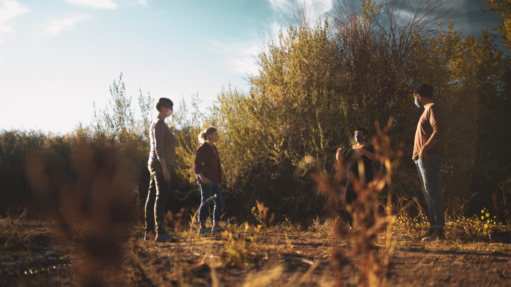 Low view of four people in facemasks talking to each over in a woodsy area.