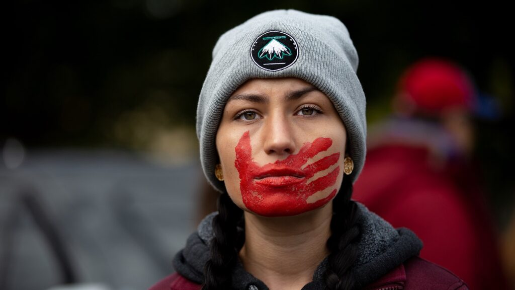 A demonstrator wearing a beanie depicting four feathers, and a red handprint covering their mouth.