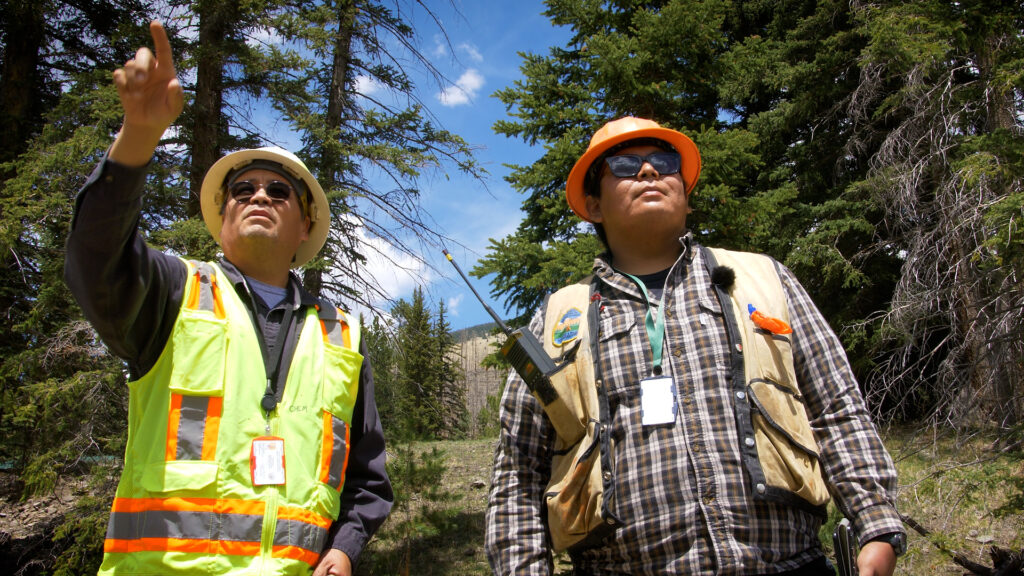 Two people in outdoor safety gear watch and discuss something off-frame.