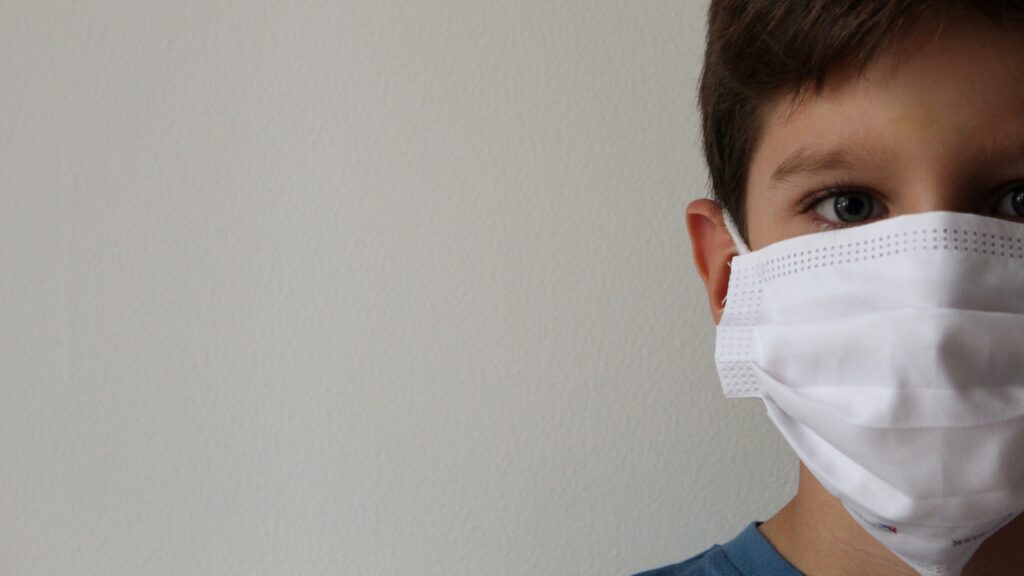 A child stands, wearing a white facemask.