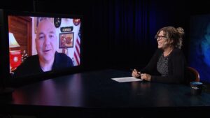 A person sitting at a table conducting an interview over television.