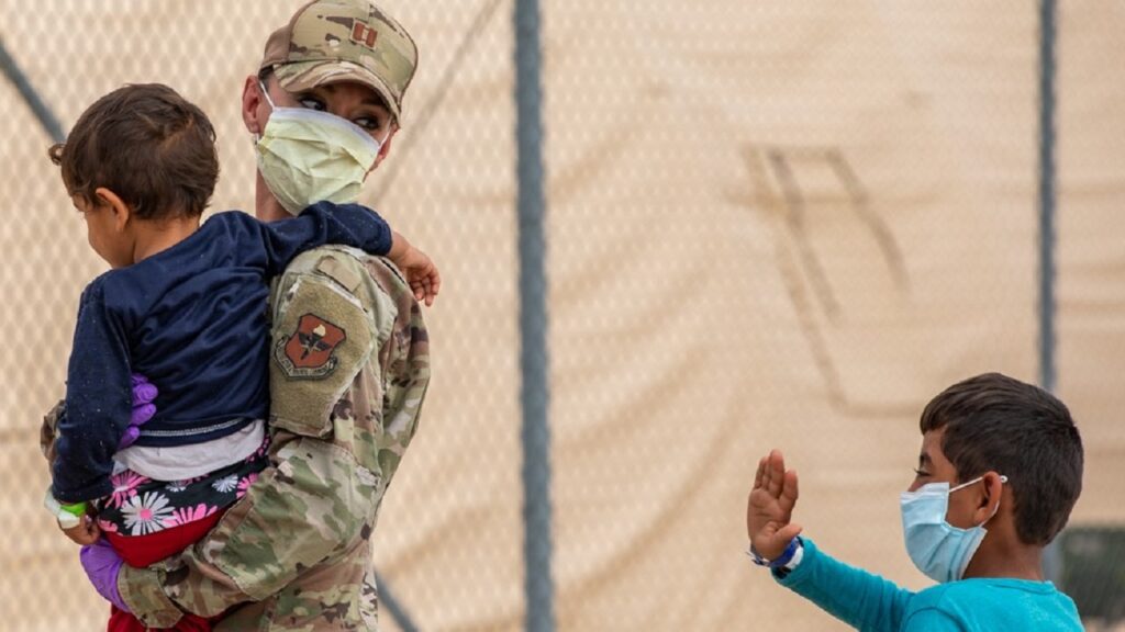 An American soldier wearing a facemask carries an Afghan toddler, with another child walking next to them.