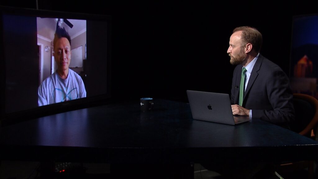 A man with a laptop speaks with someone over videochat on a television.