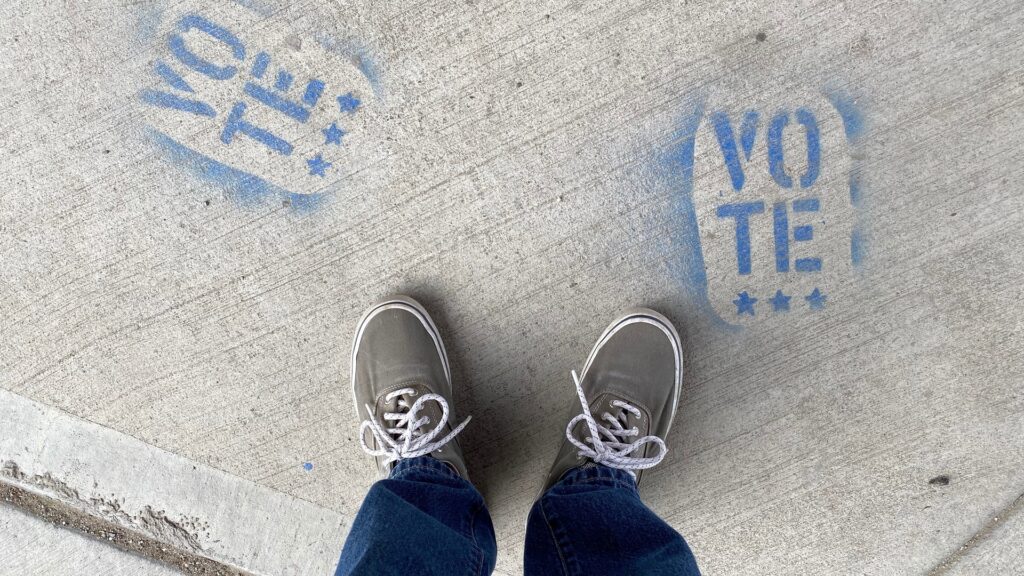 Looking down at a person's shoes standing on concrete with "Vote" spray painted on it