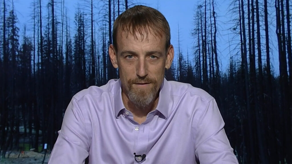 Matthew Hurteau sits for an interview in front of screen displaying a burnt forest.