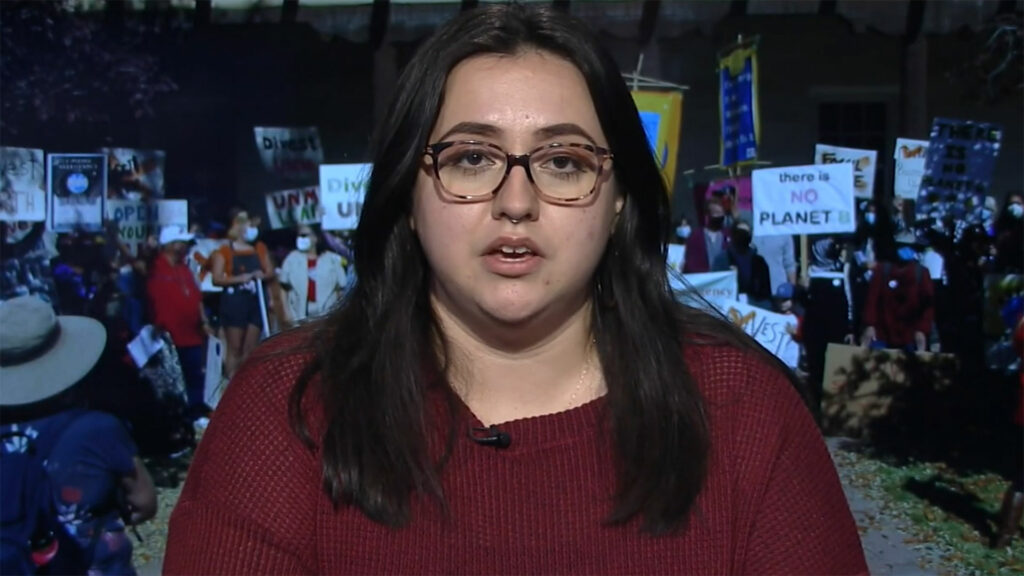 Sofia Jenkins-Nieto sits for an interview in front of a screen displaying the divest protest at UNM.