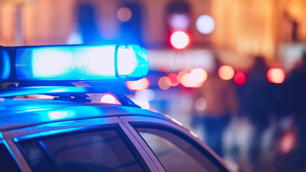 The top of a police car with the lights turned on with some out of focus people in the back.