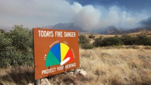 A fire danger sign indicating a high risk with a plume of smoke behind in the distance.