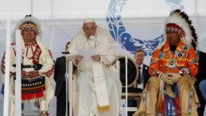 Pope Francis stands between to tribal elders in traditional outfits and delivers an apology for boarding schools.