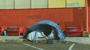 A homeless persons tent on the sidewalk in a parking lot.