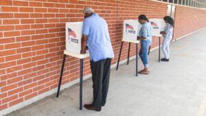 Three people lined up along a brick wall at voting tables casting their votes.