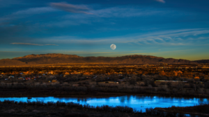 An image of the Sandia Mountains and the Rio Grande softly illuminated by the sunsetting and the moon emerging.